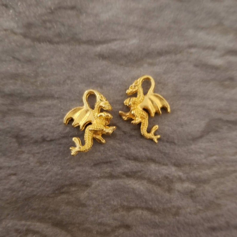 K&ouml;nnte beinhalten: Zwei goldfarbene Drachen-Anh&auml;nger mit Fl&uuml;geln und Schw&auml;nzen. Die Drachen sind in entgegengesetzte Richtungen gerichtet.