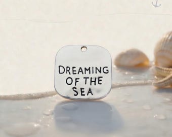 May include: A square silver-toned metal charm with rounded corners featuring the stamped black text "DREAMING OF THE SEA" and a small hole at the top for attachment. The charm has a smooth, reflective surface and is positioned upright.