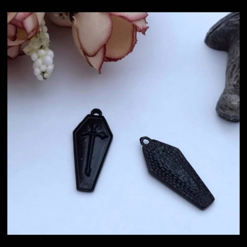 May include: Two black metal coffin charms. One charm has a cross design and the other has a textured surface.