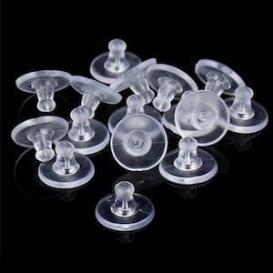 May include: A close-up of a pile of clear plastic earring backs. The backs are designed to be used with stud earrings and have a small, round, adhesive pad on the bottom.