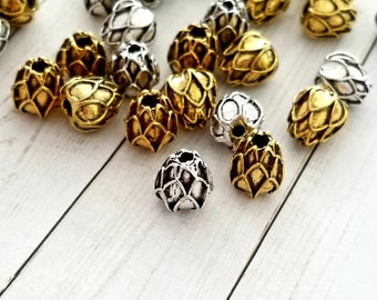 May include: A collection of gold and silver metal beads with a textured, pinecone-like design. The beads are scattered on a white wooden surface.