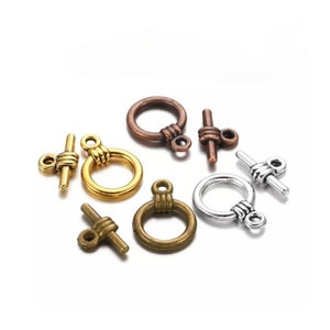 May include: Assortment of five metal toggle clasps in gold, silver, and copper finishes. Each clasp features a round ring and a T-shaped bar with a decorative knot.