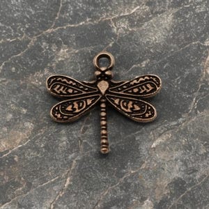 May include: A bronze-colored dragonfly charm with intricate wing detailing. The dragonfly has a loop at the top for attaching to jewelry or other crafts. The wings feature a detailed pattern, and the body is segmented.