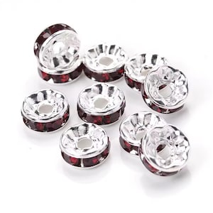 May include: A collection of silver-colored, round beads with a central hole. Each bead is accented with a ring of small, dark red gemstones. The beads are scattered on a white surface, creating a sparkling effect.
