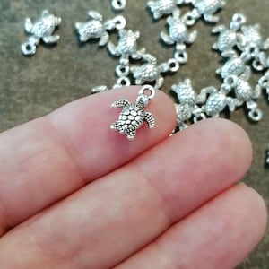 May include: A close-up of a silver turtle charm with a textured shell. The charm has a loop at the top for attaching to a necklace or bracelet.