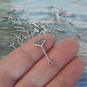 May include: A silver metal rake charm with a loop for attaching to a necklace or bracelet. The charm is small and detailed, with a realistic rake design.
