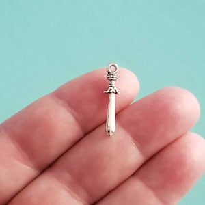 May include: Silver sword charm with a detailed hilt and a pointed tip. The charm is small and delicate, perfect for adding a touch of fantasy to jewelry or crafts.