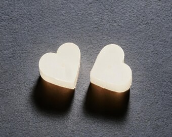 May include: Two heart-shaped, off-white objects sit on a dark gray surface. The objects have a soft, glowing appearance, with subtle shadows suggesting depth. The hearts are smooth and solid, with rounded edges.