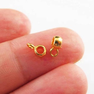 May include: Two small, gold-colored jewelry findings. One is a round bail with a small loop, and the other is a cylindrical bead bail with a loop. Both are shiny and appear to be made of metal.