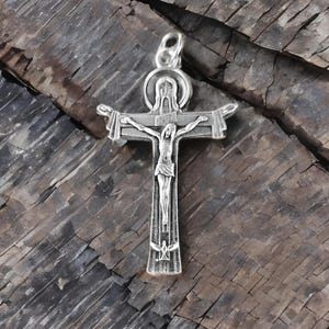 May include: A silver-colored crucifix pendant with a detailed depiction of Jesus Christ on the cross. The cross features a halo above the head and the inscription "INRI". The pendant is displayed on a rustic, weathered wooden surface.