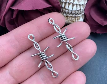 May include: Two silver-colored barbed wire charms. Each charm has a twisted wire design with horizontal barbs. The charms are displayed against a background of a hand, a skull, and dark red flowers.