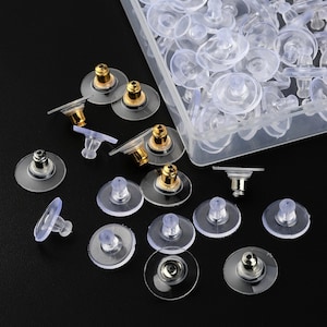 May include: A collection of earring backs featuring clear, flexible silicone discs and metallic gold or silver-toned metal inserts. Some backs are loose, while others are contained within a clear, rectangular plastic storage container.