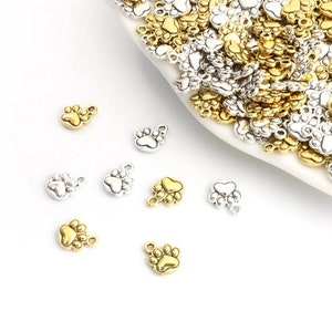 May include: A collection of gold and silver paw print charms. The charms are small and have a loop at the top for attaching to jewelry or other crafts.