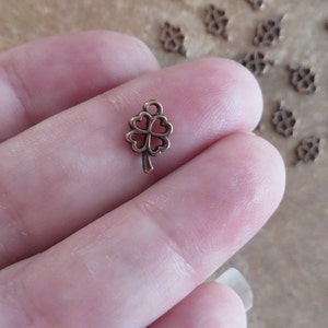 May include: A close-up of a small, copper-colored four-leaf clover charm. The charm has a loop at the top for attaching to a necklace or bracelet.