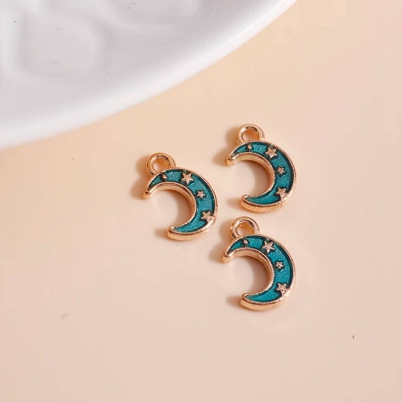 Op de afbeelding: Drie goudkleurige halve maan bedels met turquoise emaille en ster details. Elke bedel heeft een kleine lus aan de bovenkant om aan sieraden te bevestigen. De bedels worden gepresenteerd op een licht perzik oppervlak.