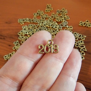 May include: A close-up of a gold-colored metal charm with the year 2018 embossed on it. The charm is being held by a hand, and there are many other similar charms scattered around it.
