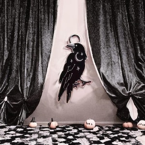 May include: A black raven-shaped pendant with a crescent moon and star details. The pendant hangs against a light background, with black curtains on either side. Small pumpkins are arranged on a black and white checkered surface.