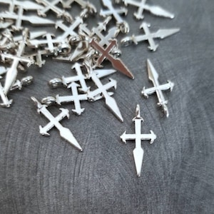 May include: A pile of silver-toned metal charms shaped like a cross with a pointed tip. The charms are scattered on a gray surface.