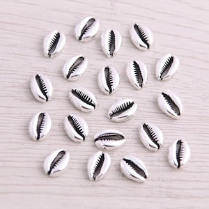 May include: A collection of silver-colored cowrie shell beads. Each bead has a detailed, textured design and a central opening. The beads are scattered on a light-colored wooden surface, creating a beachy, nautical aesthetic.