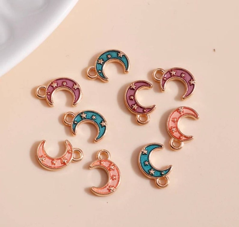 Op de afbeelding: Een verzameling maanvormige bedels in verschillende kleuren, waaronder roze, blauwgroen en paars, met goudkleurige randen. Elke bedel heeft kleine sterren en een lus om aan sieraden of andere knutselwerken te bevestigen.