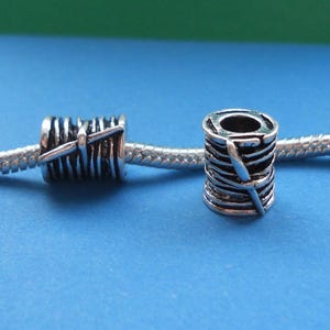 May include: Two silver-colored charm beads, each designed to resemble a spool of thread with a needle. The beads are cylindrical with detailed grooves and a needle design wrapped around them. They are displayed on a silver chain against a blue background.