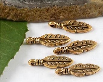 May include: Close-up of five small, gold-colored leaf charms. Each charm has a detailed leaf design with a small loop at the top for stringing. The charms are arranged on a white surface, with a green leaf and textured background visible.