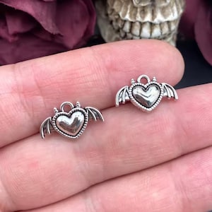 May include: Two small, silver-toned heart charms with bat wings and tiny horns. The charms are detailed with a textured border and a loop at the top for attaching to jewelry. The background includes a skull and dark red flowers.
