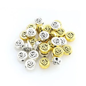 May include: A pile of round metal beads with the Om symbol in a raised design. The beads are a mix of silver and gold.