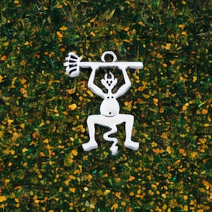 May include: A silver-colored pendant featuring a stylized figure lifting a barbell overhead. The figure has a circular head, a small dot for a belly button, and zig-zag legs. The barbell has a textured end. The pendant is set against a green and yellow background.