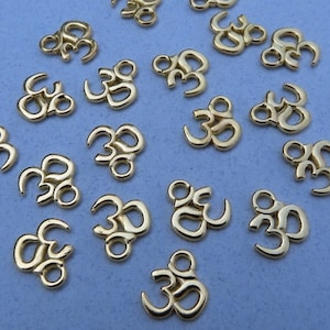 May include: A collection of gold-colored Om symbol charms. Each charm features the stylized Sanskrit symbol, with a loop at the top for attachment. The charms are scattered on a light blue surface.