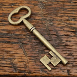 May include: An antique-style brass key featuring a rounded bow, a slender cylindrical shaft with a single decorative ring, and a bit with a distinct geometric cutout pattern. The metal surface displays a muted, aged gold-toned finish.