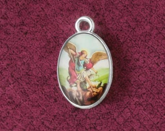 May include: Oval pendant with a silver-colored frame. The pendant features a colorful illustration of an angel with a sword, battling a creature. The background is a light green and blue. The pendant has a loop at the top for a chain.