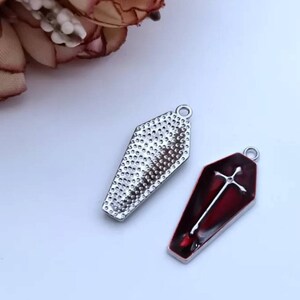 Op de afbeelding: Twee zilveren kistcharms, een met een rode emaille afwerking en een wit kruisontwerp, de andere met een getextureerd oppervlak.