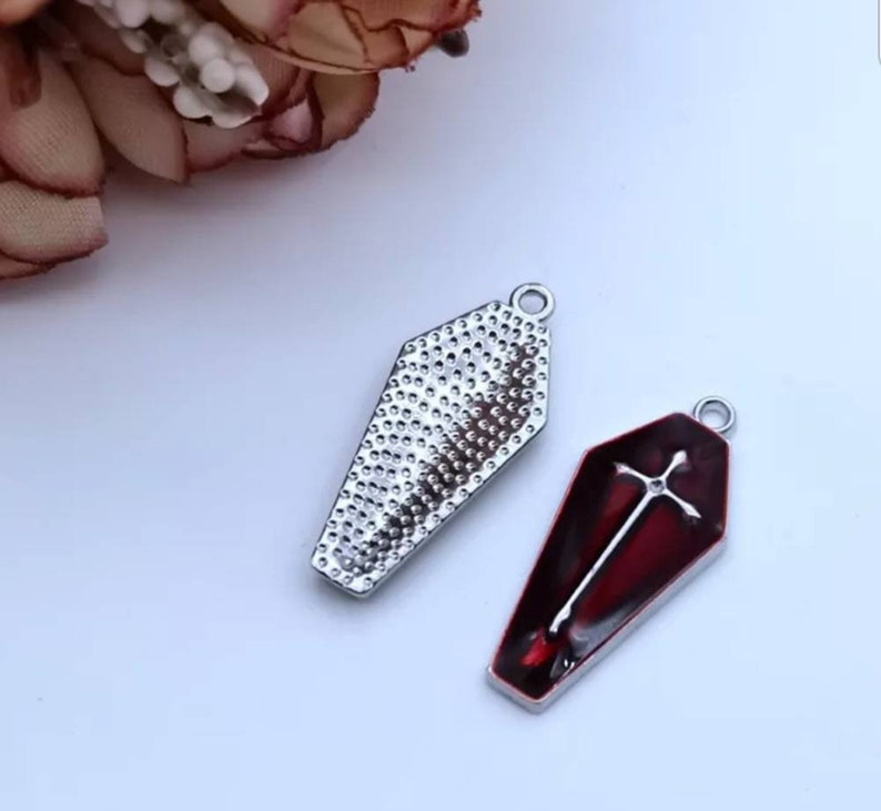 Op de afbeelding: Twee zilveren kistcharms, een met een rode emaille afwerking en een wit kruisontwerp, de andere met een getextureerd oppervlak.
