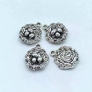 May include: Four silver-colored bird nest charms, each with a loop for attaching to jewelry. Each nest contains three small, round eggs. The charms have a detailed, textured appearance, perfect for crafting.