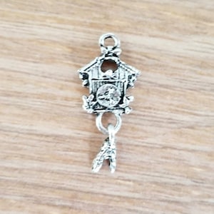 May include: A silver-colored charm in the shape of a cuckoo clock. The charm features a detailed miniature clock face, decorative elements, and a dangling pendulum. The charm is designed with a loop at the top for easy attachment to a necklace or bracelet.
