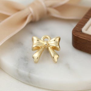 May include: A small, polished gold-tone metal charm shaped like a bow. The charm features a smooth, reflective surface with a central knot and two hanging loops, and includes a small circular jump ring at the top for attachment purposes.