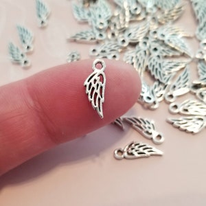 May include: A close-up of a silver angel wing charm. The charm is small and delicate, with a detailed design. It is perfect for adding a touch of elegance to jewelry or crafts.