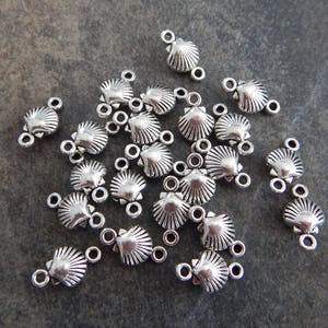May include: A group of silver-toned metal seashell charms with two jump rings on each charm. The charms are scattered on a gray textured surface.