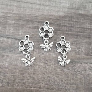 Op de afbeelding: Drie zilverkleurige bijenbedels. Elke bedel heeft een honingraatvorm met een kleine bijenhanger eronder. De bedels worden gepresenteerd op een grijze houten ondergrond. Geschikt voor het maken van sieraden of knutselprojecten.