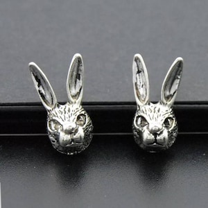 May include: A pair of silver-colored stud earrings, each shaped like a rabbit's head. The rabbit heads have detailed features, including long ears and facial features. The earrings are displayed on a dark surface.
