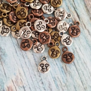 May include: A pile of silver, gold, and copper metal charms shaped like faces with different expressions. The charms have a loop at the top for attaching to jewelry or other crafts.