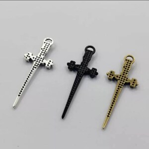 May include: Three silver, black, and gold sword-shaped cross charms. The charms are detailed with a textured surface and have a loop at the top for attaching to jewelry.