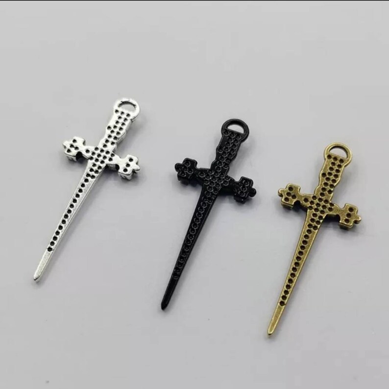 May include: Three silver, black, and gold sword-shaped cross charms. The charms are detailed with a textured surface and have a loop at the top for attaching to jewelry.