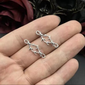 May include: Two silver-toned metal charms shaped like barbed wire. The charms are in the shape of a loop with two barbed wire strands on each side.