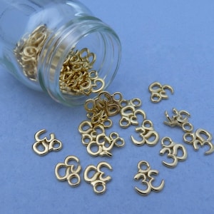 May include: A clear glass jar tilted on its side, spilling out numerous small, gold-colored metal charms shaped like the Om symbol. Each charm features a small loop at the top for attachment, and they are scattered across a flat, light blue surface.