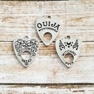 May include: Three silver heart-shaped charms with a hole in the center. The charms have black lettering that reads "Ouija Mystifying Oracle", "Yes No Goodbye Hello", and "Witching Hour".