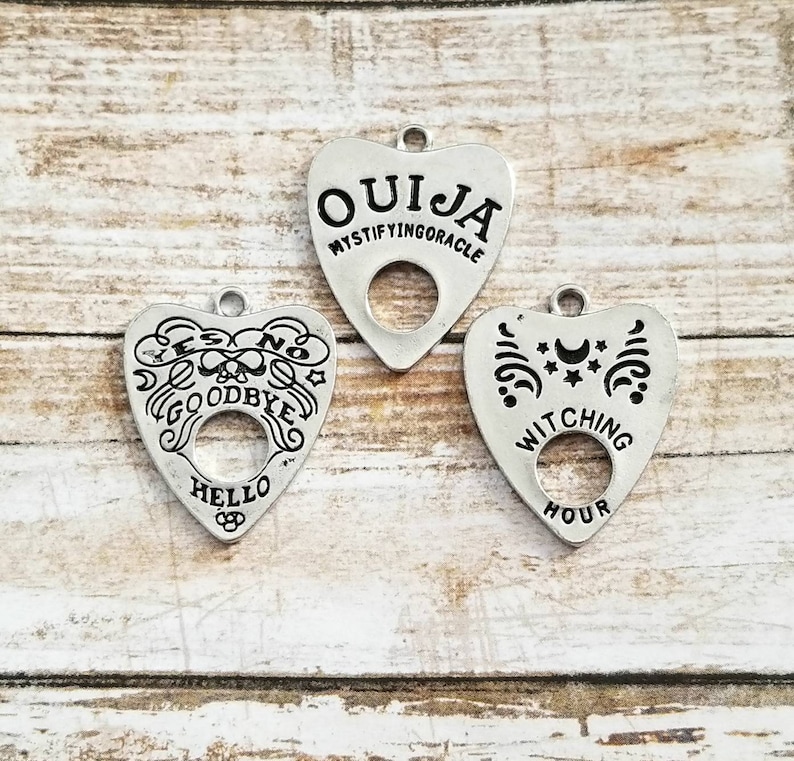 May include: Three silver heart-shaped charms with a hole in the center. The charms have black lettering that reads "Ouija Mystifying Oracle", "Yes No Goodbye Hello", and "Witching Hour".