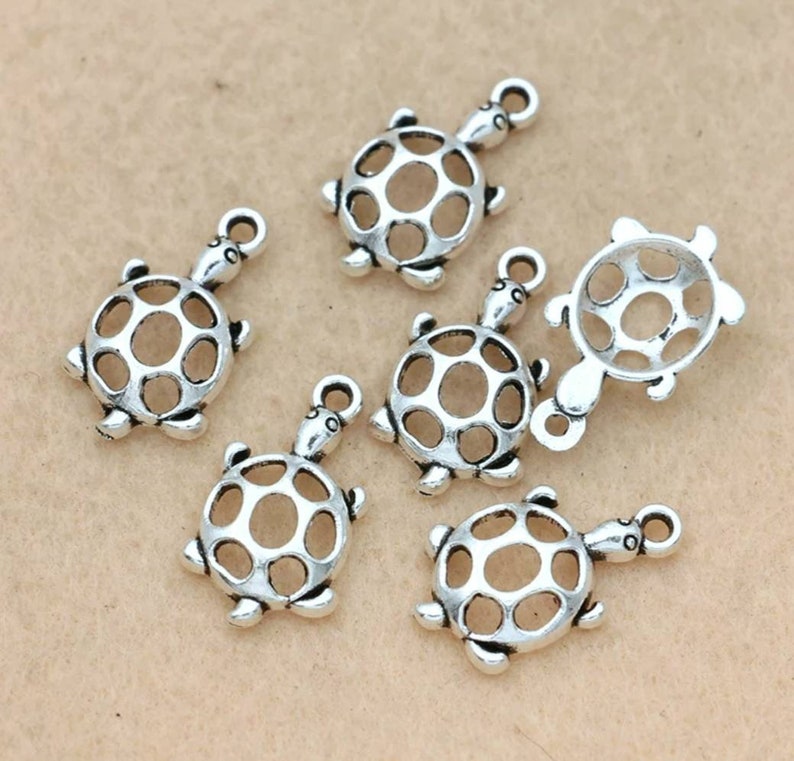 Puede incluir: Un conjunto de abalorios plateados con forma de tortuga. Cada abalorio presenta un dise&ntilde;o hueco y calado en el caparaz&oacute;n y un lazo para sujetarlo a joyas u otras manualidades. Los abalorios son peque&ntilde;os, con una cabeza y patas de tortuga detalladas.