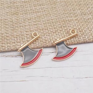 May include: Two gold-colored axe charms with gray blades and red trim. The axes have a loop at the top for attaching to jewelry or other crafts. The background is a light tan fabric.